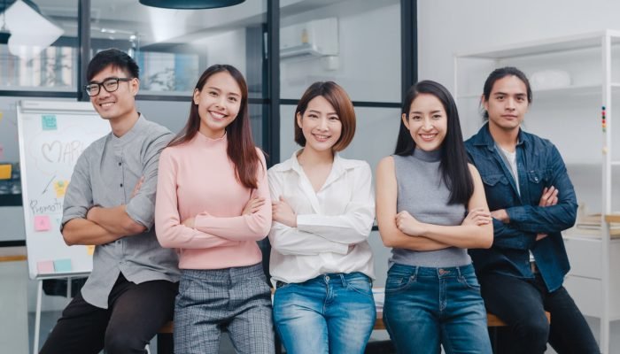 Group of Asia young creative people in smart casual wear smiling and arms crossed in creative office workplace. Diverse Asian male and female stand together at startup. Coworker teamwork concept.